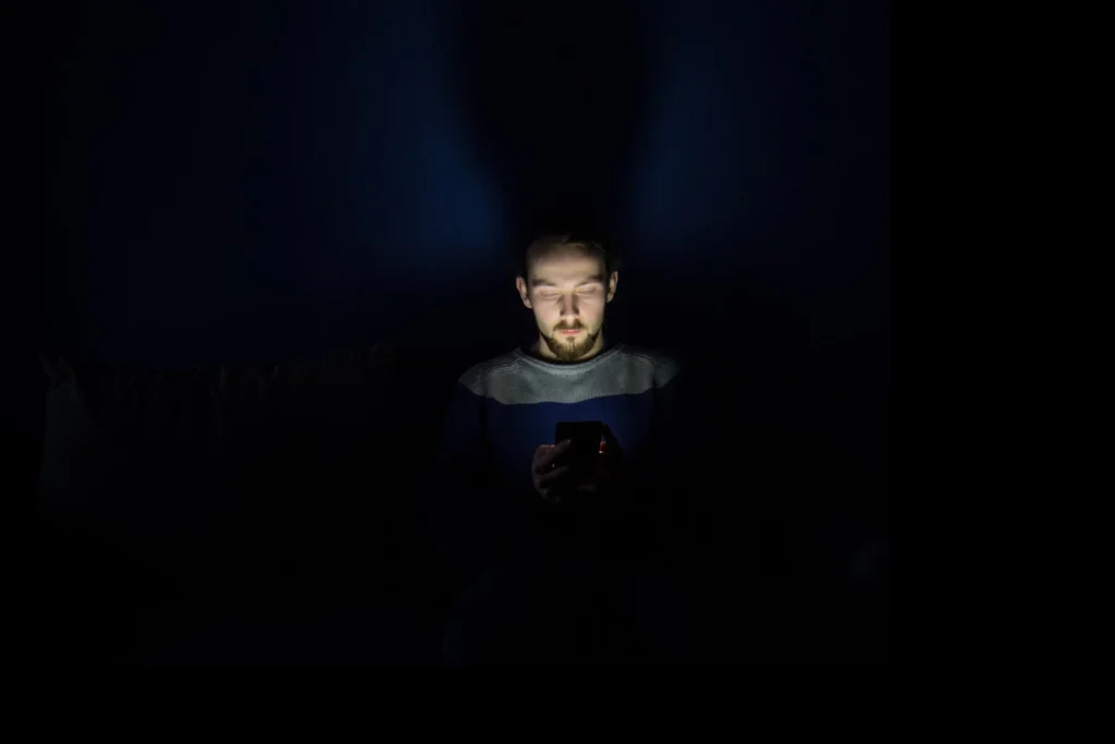 A man sitting in a dark room illuminated by his smartphone screen, representing phone addiction and late-night scrolling